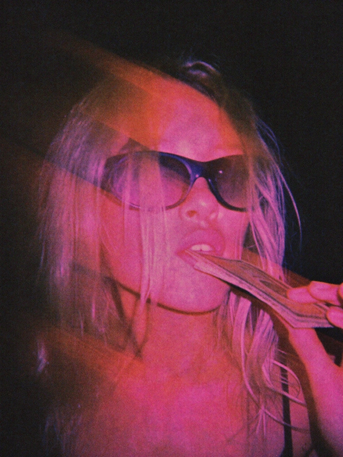 Raw motion-blurred flash photo of a woman in sunglasses biting a stack of cash, saturated pink and magenta tones